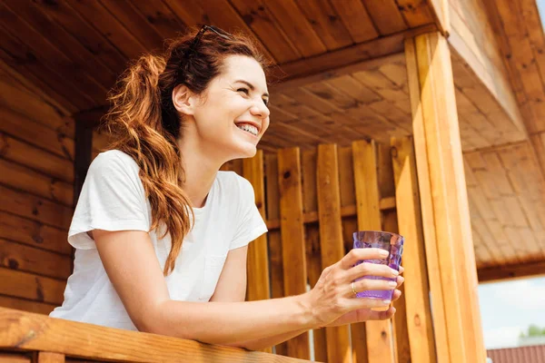 Jeune femme souriante avec verre de boisson debout sur la terrasse du chalet en bois et regardant loin — Photo de stock