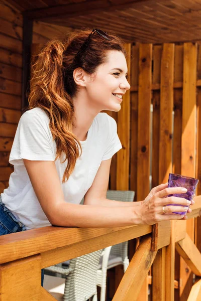 Felice giovane donna con bicchiere di bevanda in piedi sulla terrazza del cottage in legno e guardando altrove — Foto stock