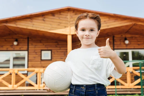 Прелестный маленький ребенок с волейбольным мячом, показывающий большой палец и смотрящий в камеру — стоковое фото