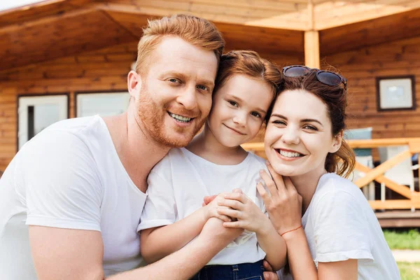 Close-up shot of beautiful young family embracing in front of wooden cottage and looking at camera — Stock Photo