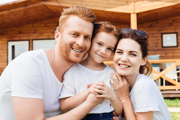 Nahaufnahme einer schönen jungen Familie, die sich vor einem Holzhaus umarmt und in die Kamera blickt — Stockfoto