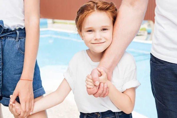 Niña feliz cogida de la mano de los padres y mirando a la cámara delante de la piscina - foto de stock