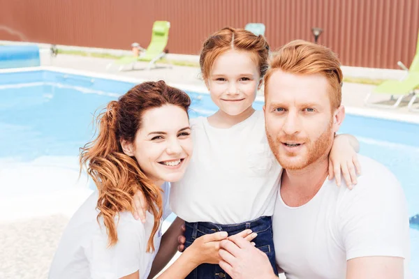 Hermosa familia joven abrazándose frente a la piscina y mirando a la cámara - foto de stock