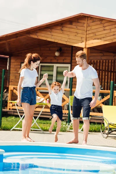 Bela jovem família jogando na beira da piscina de casa de campo de madeira aconchegante — Fotografia de Stock