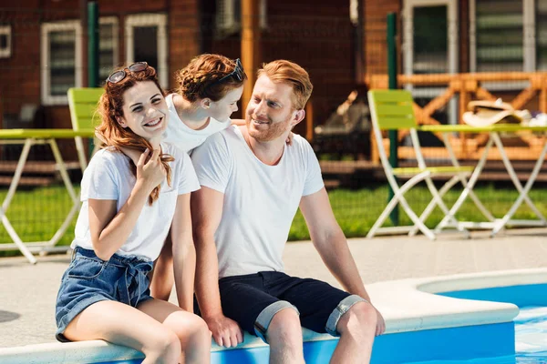 Bela jovem família sentada à beira da piscina e abraçando juntos — Fotografia de Stock