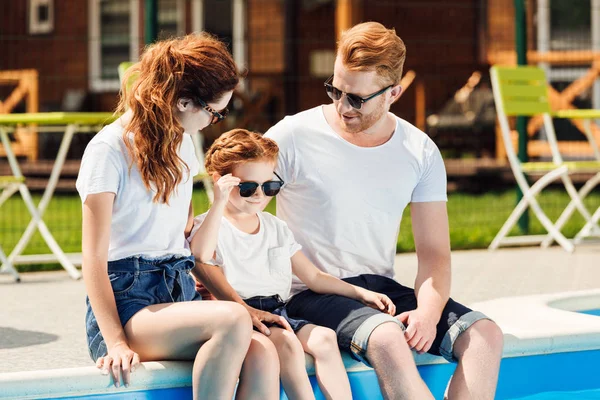 Familia joven en camisetas blancas y gafas de sol sentados en la piscina juntos - foto de stock