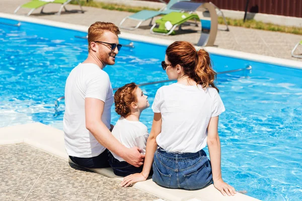 Happy young family in white t-shirts and sunglasses sitting on poolside together — Stock Photo
