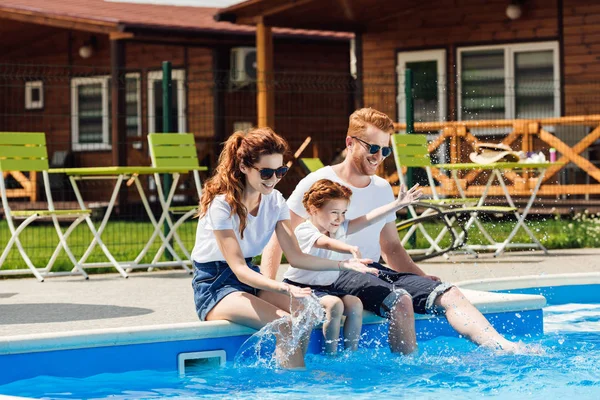 Bella giovane famiglia in t-shirt bianche e occhiali da sole seduti a bordo piscina insieme — Foto stock