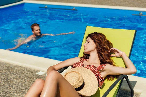 Jeune femme se détendre sur une chaise longue tandis que son petit ami nage dans la piscine sur fond — Photo de stock