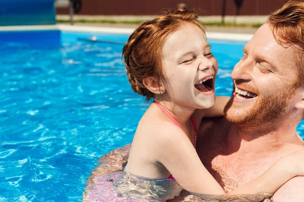 Padre riendo abrazando a su pequeña hija sonriente en la piscina - foto de stock