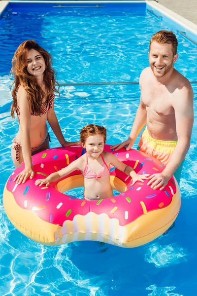 Sorridenti giovani genitori con figlia galleggiante in anello di gomma guardando la fotocamera in piscina — Foto stock