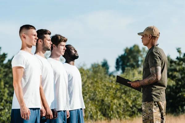 Side view of african american tactical instructor with notepad and multiracial young soldiers on range — Stock Photo