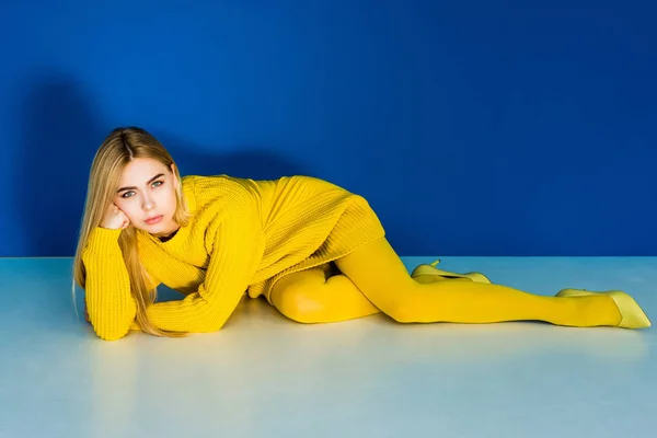 Belle fille blonde vêtue de jaune couchée sur le sol sur fond bleu — Photo de stock