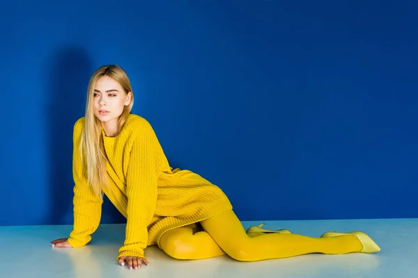 Jovem sedutora em roupas de moda amarela deitada no fundo azul — Fotografia de Stock