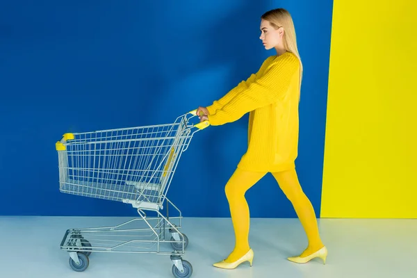 Elegante blonde Frau schiebt Einkaufswagen auf blauem und gelbem Hintergrund — Stockfoto