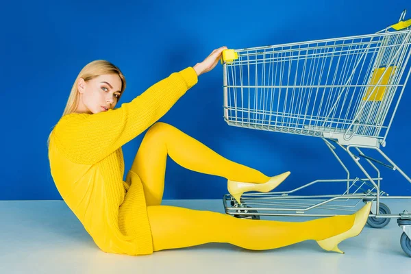 Muchacha joven atractiva vestida de amarillo sentado por el carrito de la compra sobre fondo azul - foto de stock