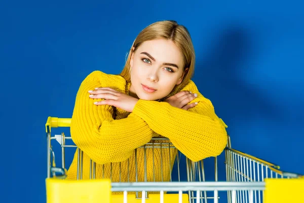 Elegant blonde woman in yellow garment leaning on shopping cart on blue background — Stock Photo