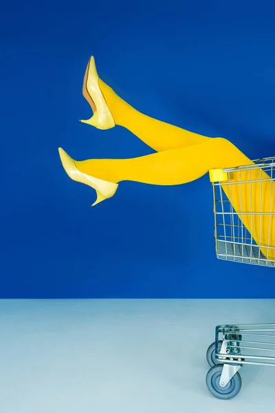 Vista cortada das pernas femininas no carrinho de compras no fundo azul — Fotografia de Stock