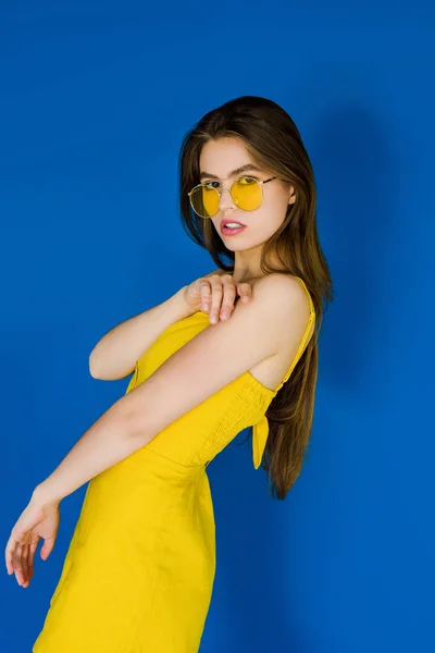 Brünette Frau in gelbem Kleid und Sonnenbrille auf blauem Hintergrund — Stockfoto