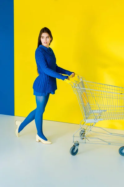 Attractive young girl with shopping cart on blue and yellow background — Stock Photo