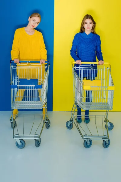 Красивые девушки в модной одежде толкают тележки для покупок на синем и желтом фоне — стоковое фото