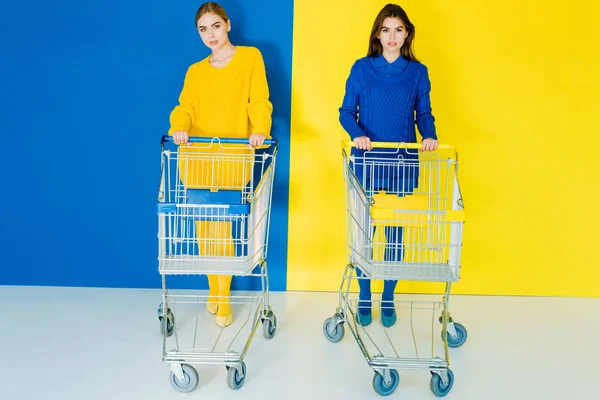 Attraktive junge Mädchen in modischer Kleidung schieben Einkaufswagen auf blauem und gelbem Hintergrund — Stockfoto