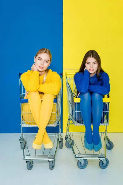 Female fashion models sitting in shopping carts on blue and yellow background — Stock Photo