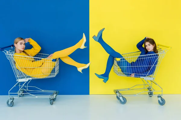 Hermosa morena y chicas rubias cabalgando en carritos de compras sobre fondo azul y amarillo - foto de stock