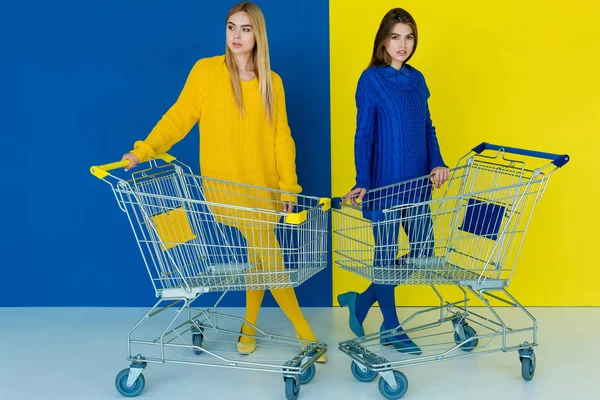 Beautiful brunette and blonde girls posing by shopping carts on blue and yellow background — Stock Photo