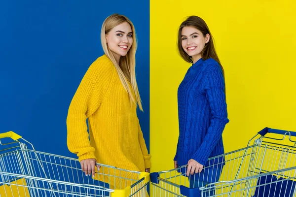 Привлекательные молодые девушки, держащие тележки с покупками и улыбающиеся изолированные на синем и желтом фоне — стоковое фото