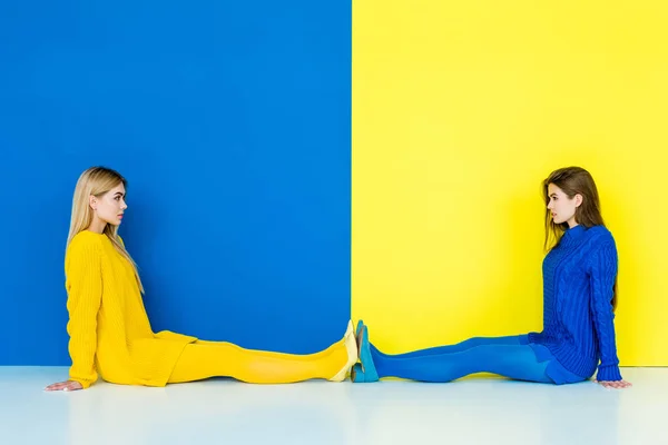 Female fashion models sitting on floor towards each other on blue and yellow background — Stock Photo