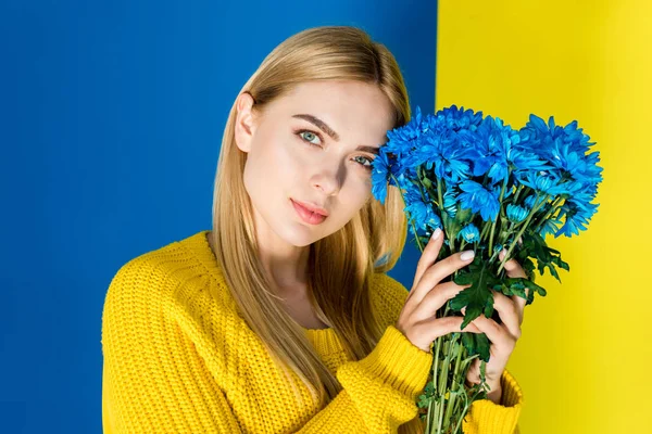 Приваблива молода дівчина в жовтому светрі тримає сині квіти ізольовані на синьо-жовтому тлі — стокове фото
