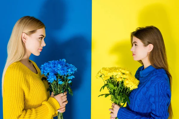 Modelos de moda femenina sosteniendo ramos de crisantemo y mirándose unos a otros sobre fondo azul y amarillo - foto de stock