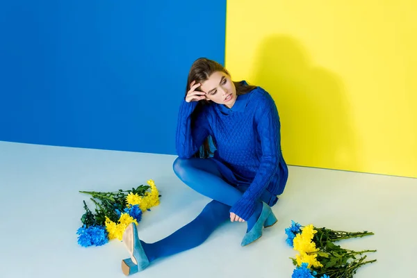 Frau in blauer Kleidung sitzt zwischen Blumen auf blauem und gelbem Hintergrund — Stockfoto