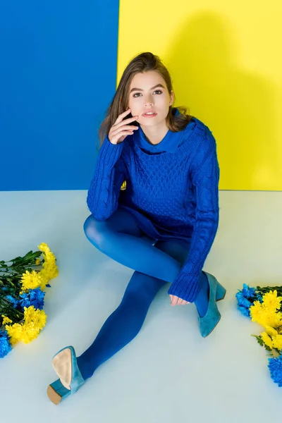 Weibliche Mode-Modell sitzt zwischen Blumen auf blauem und gelbem Hintergrund — Stockfoto
