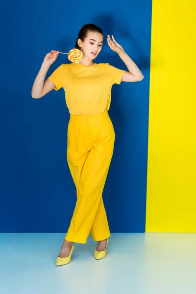 Pretty brunette girl in yellow clothes holding lollipop on blue and yellow background — Stock Photo
