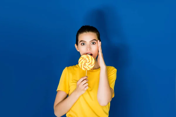 Beautiful surprised brunette girl holding lollipop on blue background — Stock Photo