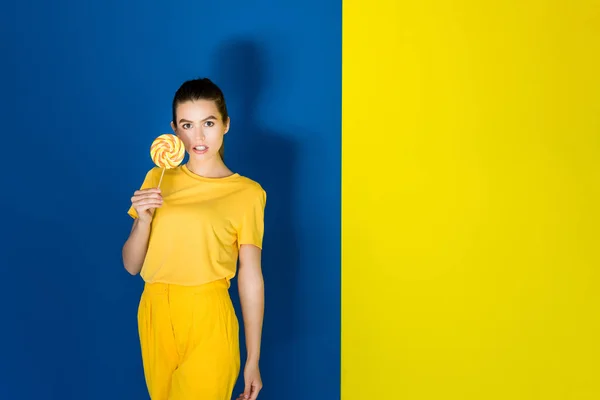 Attractive young girl in yellow outfit holding lollipop on blue and yellow background — Stock Photo