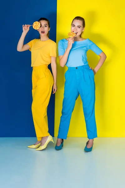 Elegantes mulheres elegantes comendo pirulitos em fundo azul e amarelo — Fotografia de Stock