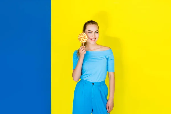 Female fashion model smiling and holding lollipop on blue and yellow background — Stock Photo