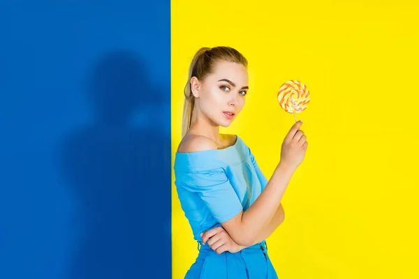 Attractive young girl holding lollipop and looking at camera on blue and yellow background — Stock Photo