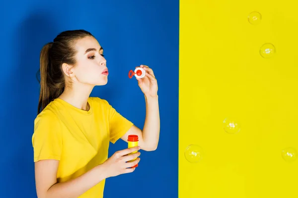 Happy young woman blowing bubbles on blue and yellow background — Stock Photo