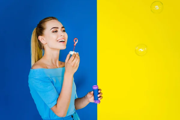 Menina atraente soprando bolhas isoladas no fundo azul e amarelo — Fotografia de Stock
