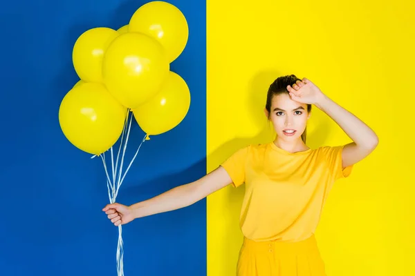 Elegant brunette woman holding bunch of yellow balloons on blue and yellow background — Stock Photo