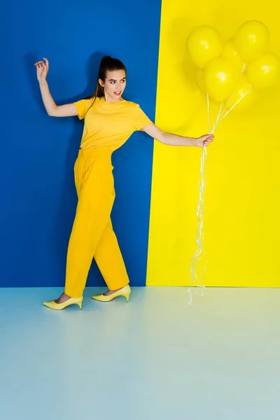 Beautiful brunette girl holding bunch of yellow balloons on blue and yellow background — Stock Photo