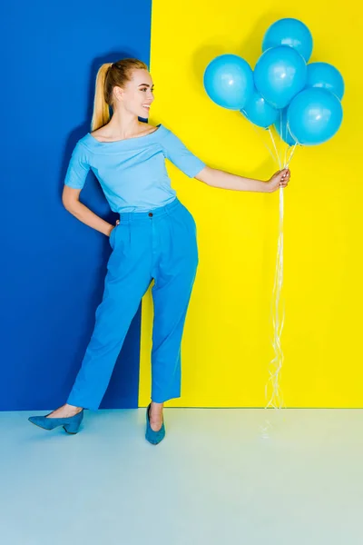 Pretty blonde woman holding bunch of blue balloons on blue and yellow background — Stock Photo