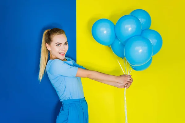 Attractive young girl holding bunch of blue balloons on blue and yellow background — Stock Photo
