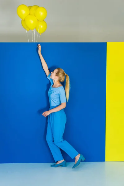 Schöne blonde Mädchen greift nach gelben Luftballons auf blauem und gelbem Hintergrund — Stockfoto