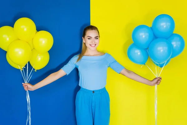 Sorridente donna bionda che tiene palloncini blu e gialli su sfondo blu e giallo — Foto stock