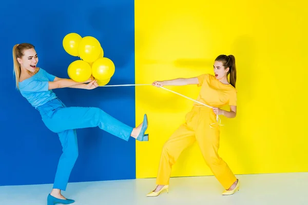 Blonde und brünette Mädchen kämpfen um Luftballons auf blauem und gelbem Hintergrund — Stockfoto